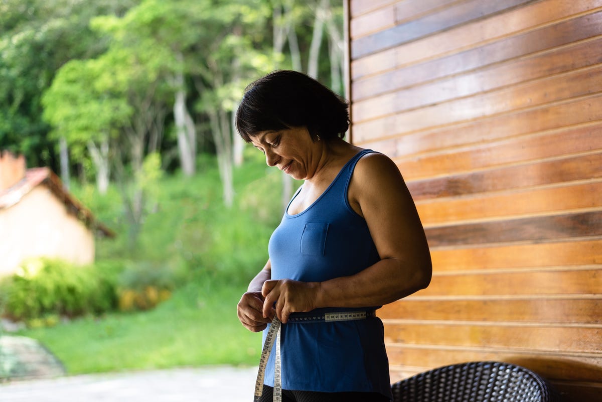 Woman in tank top measures her waist outside.