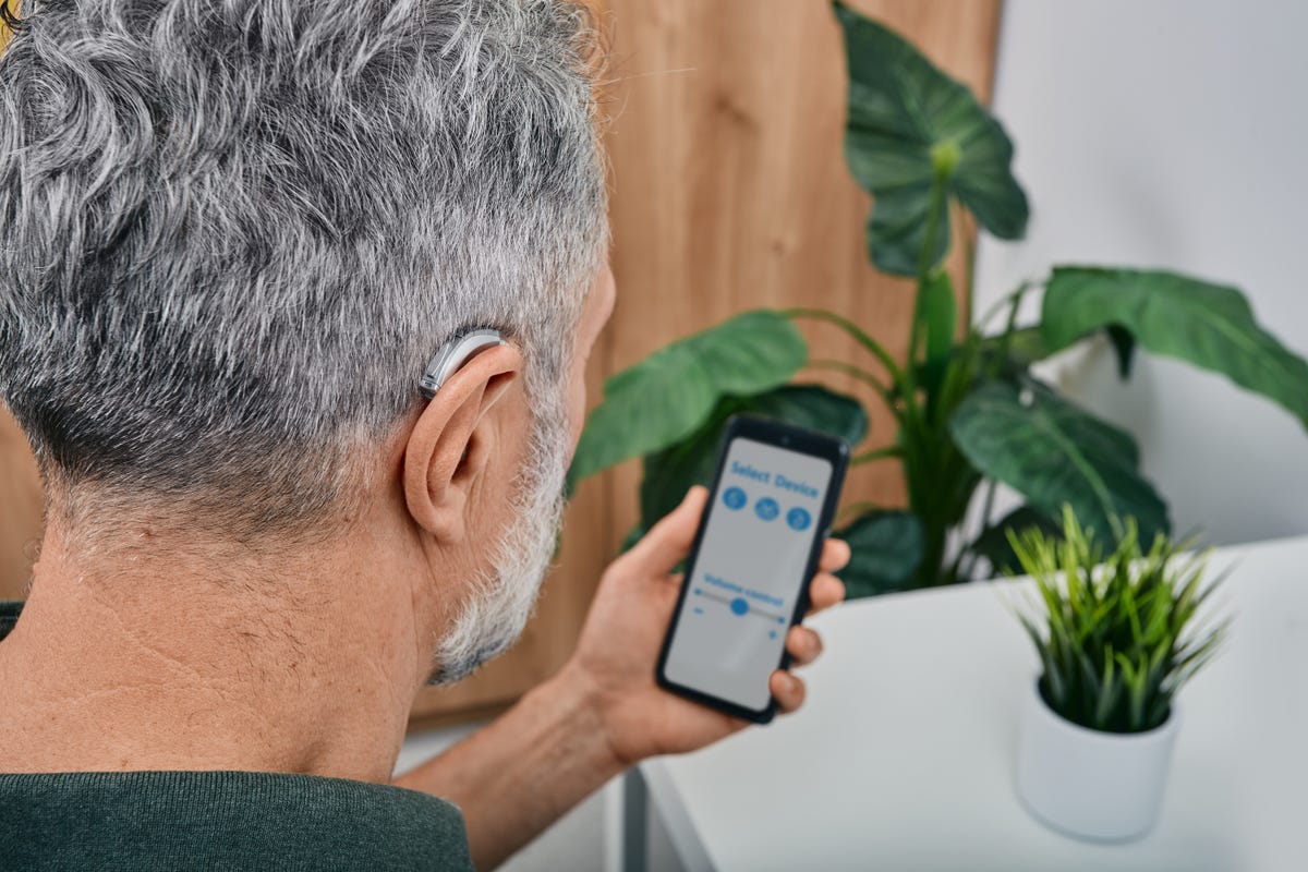 A man wearing a hearing aid adjusts at home using an app on his phone.