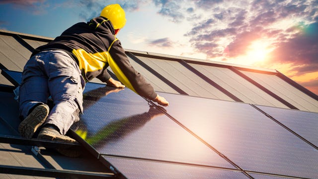 A solar panel installer places a solar panel on top of a house as the sun sets.