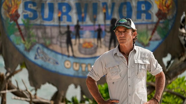 jeff probst stands with hands on hips in front of Survivor sign