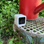 A small Wyze camera on outside plant stand next to a red watering can