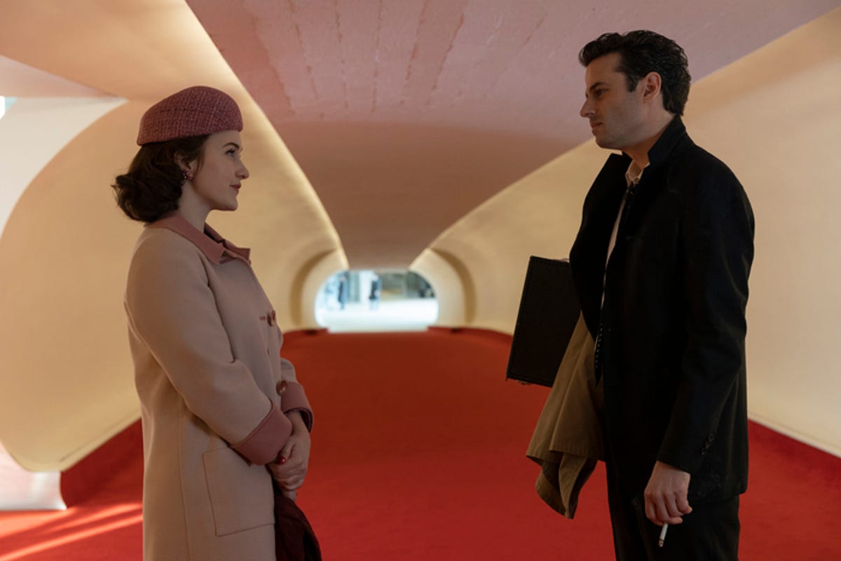 woman in coat and hat faces man in jacket as they talk in long carpeted hallway